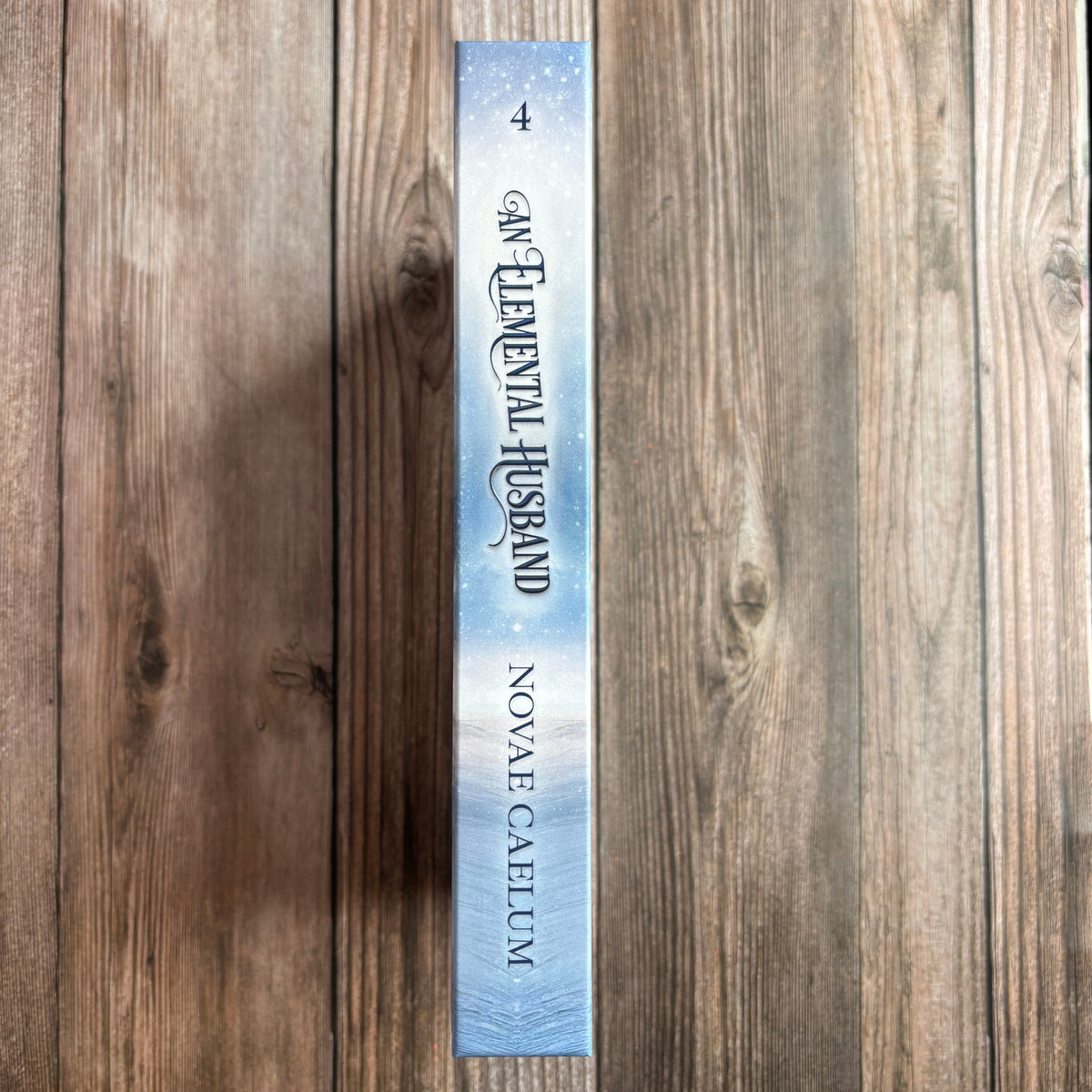 Spine of the hardcover "An Elemental Husband: The King's Weaver Book 4" by Novae Caelum, lying on a wood background, showing the "4" volume number and icy blue cover design.