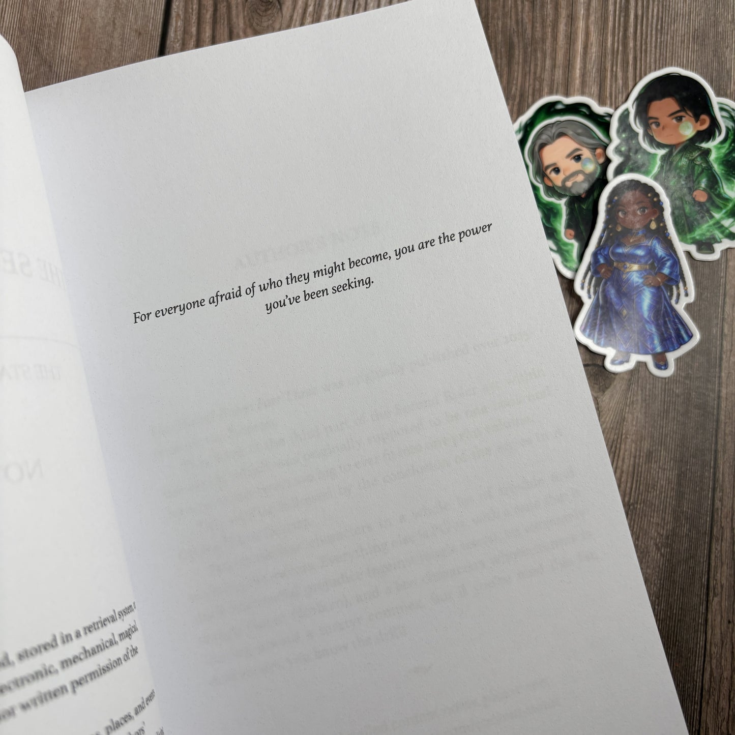 Open dedication page of "The Second Ruler: Part Three" by Novae Caelum reading "For everyone afraid of who they might become, you are the power you've been seeking," with three chibi character stickers on the wood beside the book.