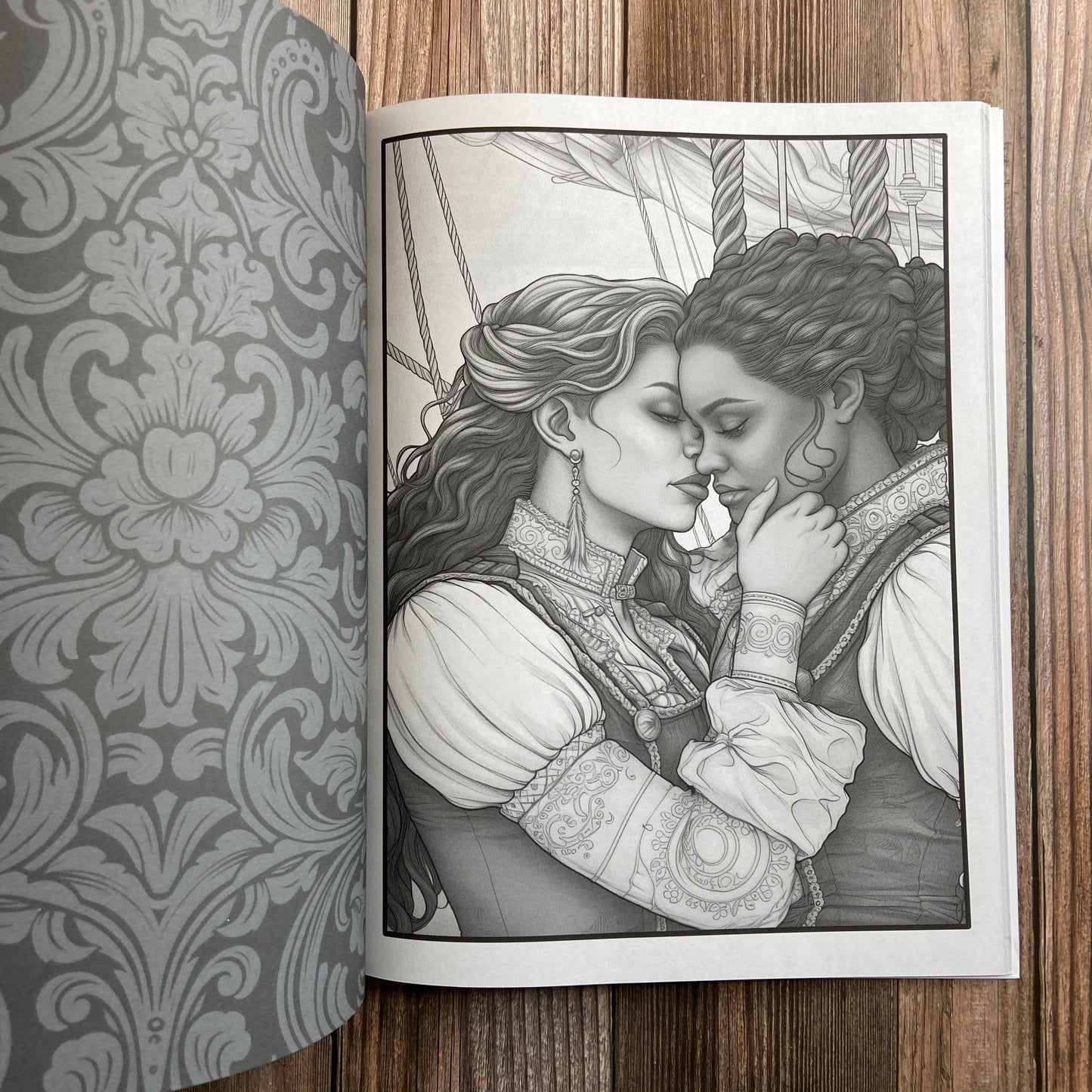 A coloring book (titled "The Sapphic Pirate Coloring Book: 30 Grayscale and Line Art Coloring Pages" by N.C. Starshadow) opened to display a black-line image of two sapphic pirates kissing.