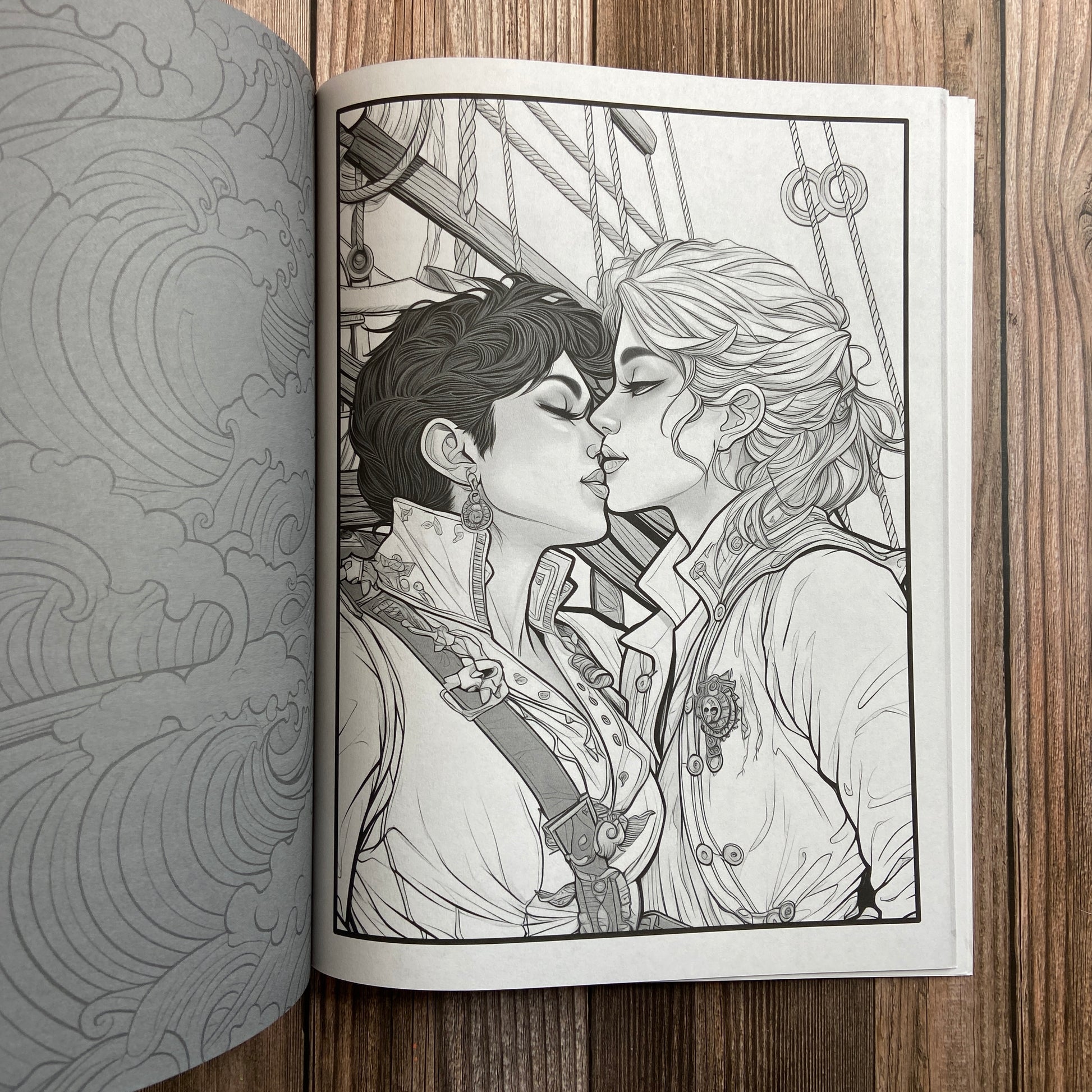 A coloring book (titled "The Sapphic Pirate Coloring Book: 30 Grayscale and Line Art Coloring Pages" by N.C. Starshadow) opened to display a black-line image of two sapphic pirates kissing.