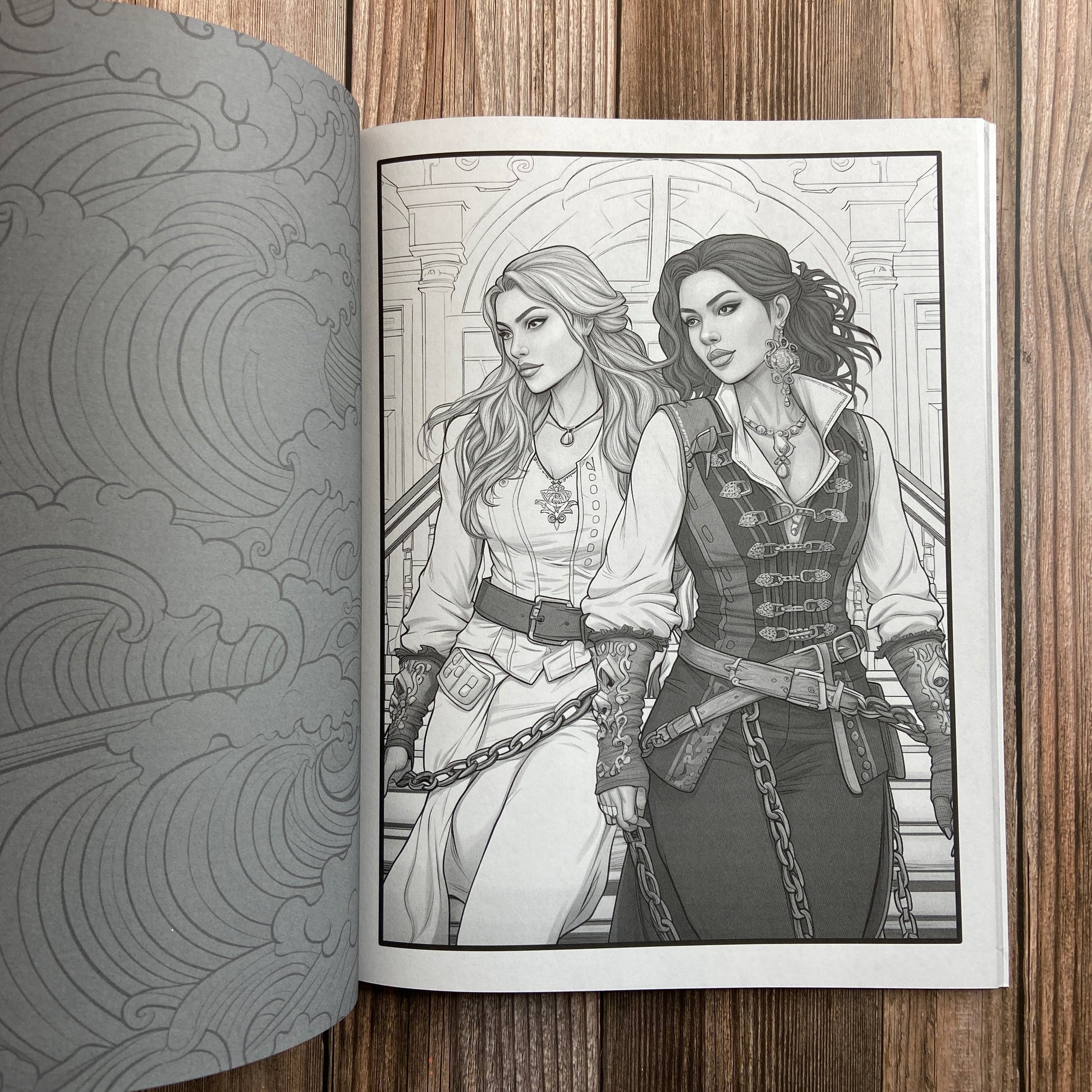 A coloring book (titled "The Sapphic Pirate Coloring Book: 30 Grayscale and Line Art Coloring Pages" by N.C. Starshadow) opened to display a black-line image of two sapphic pirates standing side-by-side.