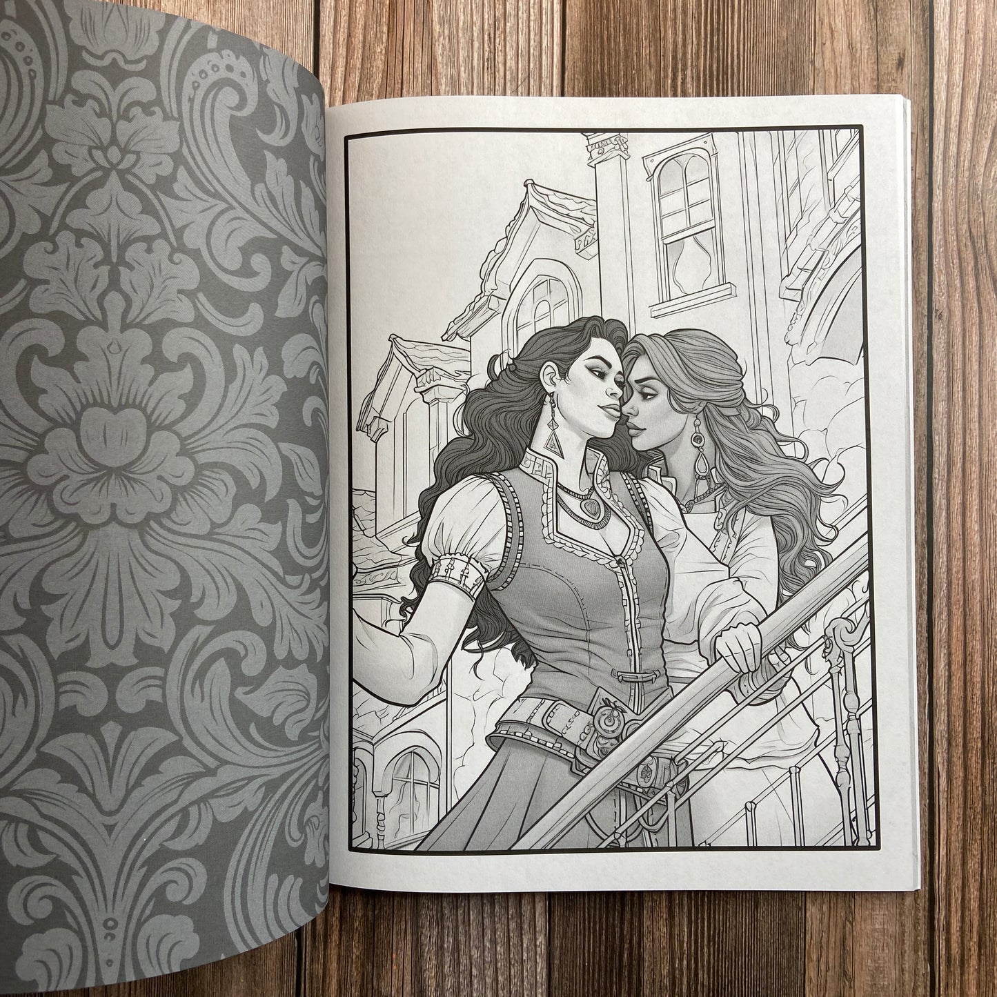 A coloring book (titled "The Sapphic Pirate Coloring Book: 30 Grayscale and Line Art Coloring Pages" by N.C. Starshadow) opened to display a black-line image of two sapphic pirates embracing.