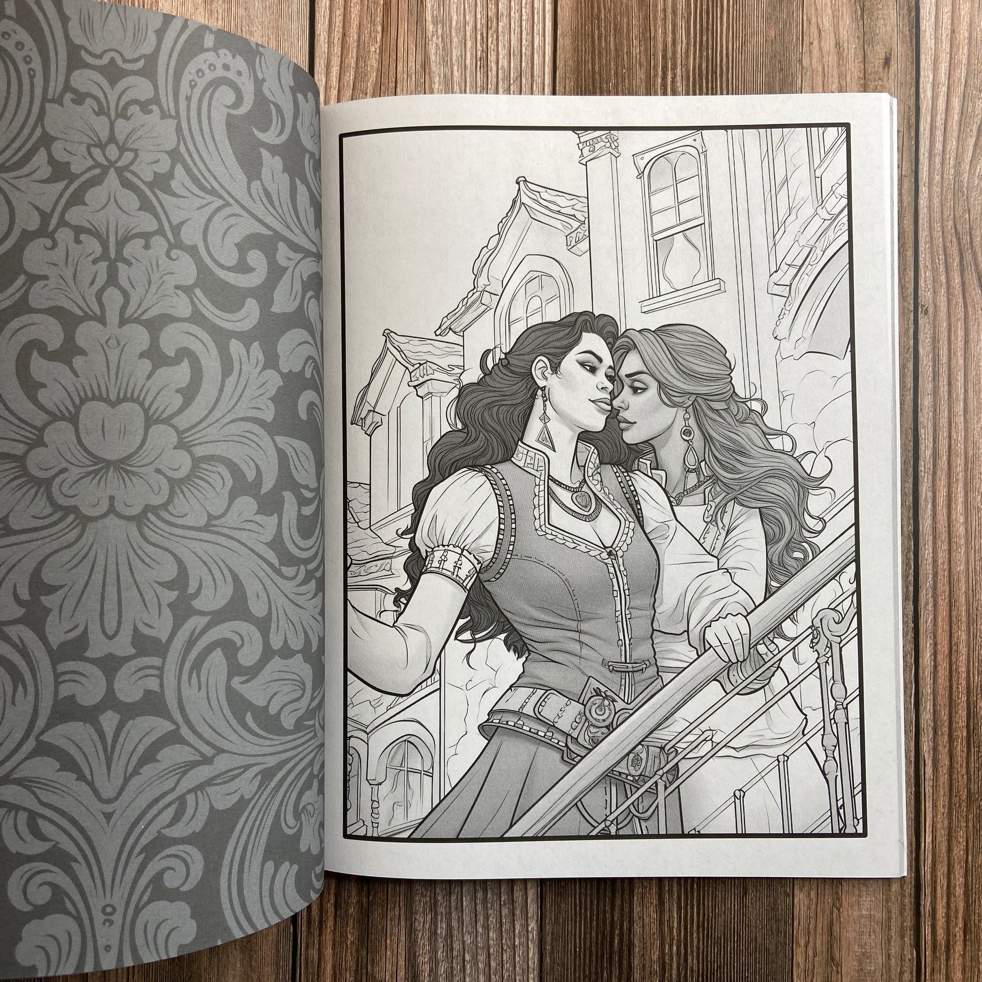 A coloring book (titled "The Sapphic Pirate Coloring Book: 30 Grayscale and Line Art Coloring Pages" by N.C. Starshadow) opened to display a black-line image of two sapphic pirates embracing.