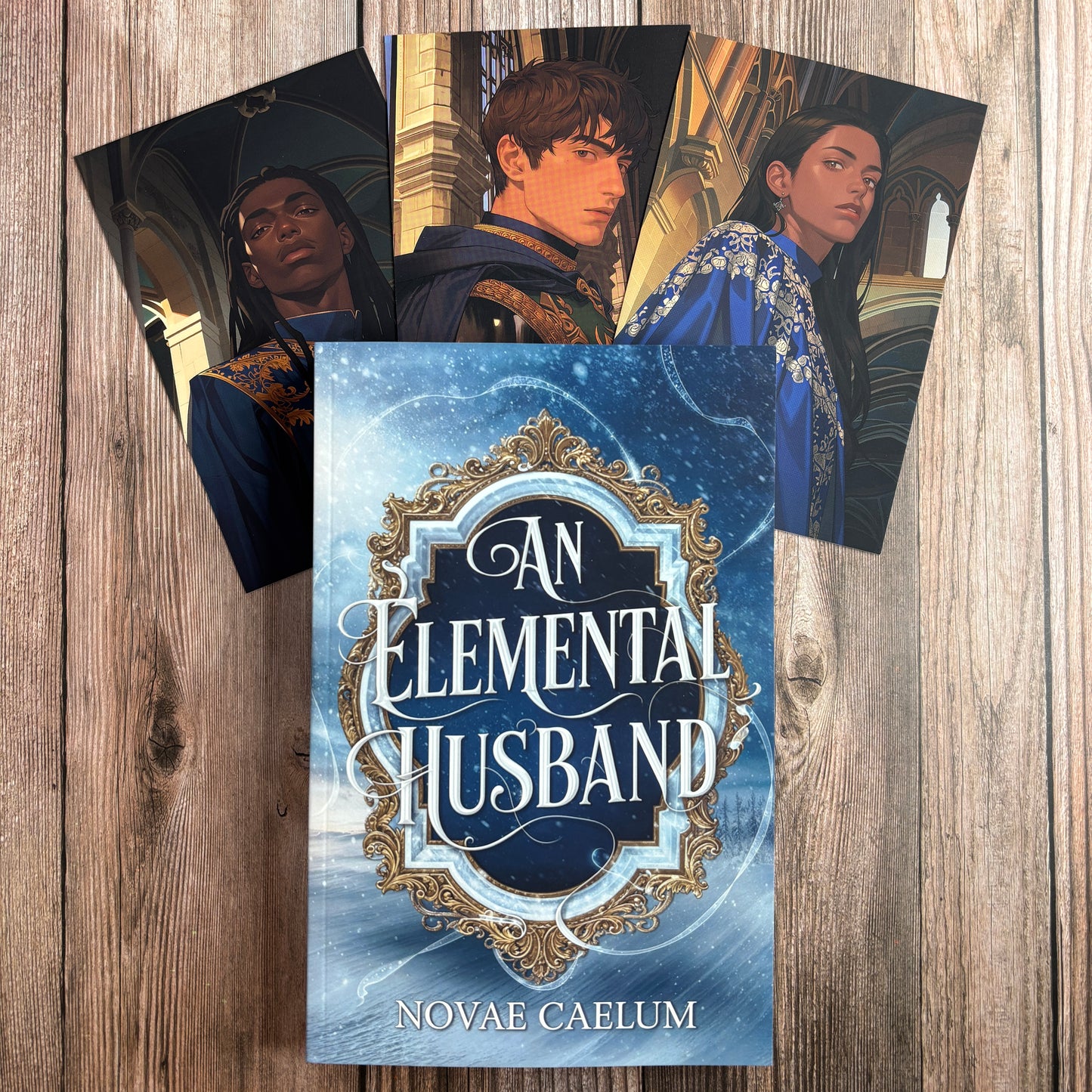 The "An Elemental Husband" paperback by Novae Caelum on a wood-plank surface, fanned behind it three character art cards: a Black man with long locs, a brown-haired man in a navy-and-gold coat, and a long-haired person in a blue-and-silver embroidered robe.