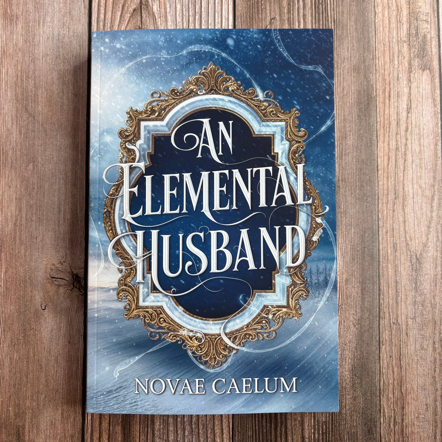 Book cover of "An Elemental Husband: The King's Weaver Book 4" by Novae Caelum, paperback edition lying flat on a wood surface, showing an ornate silver-and-gold mirror frame over a snowy winter landscape.