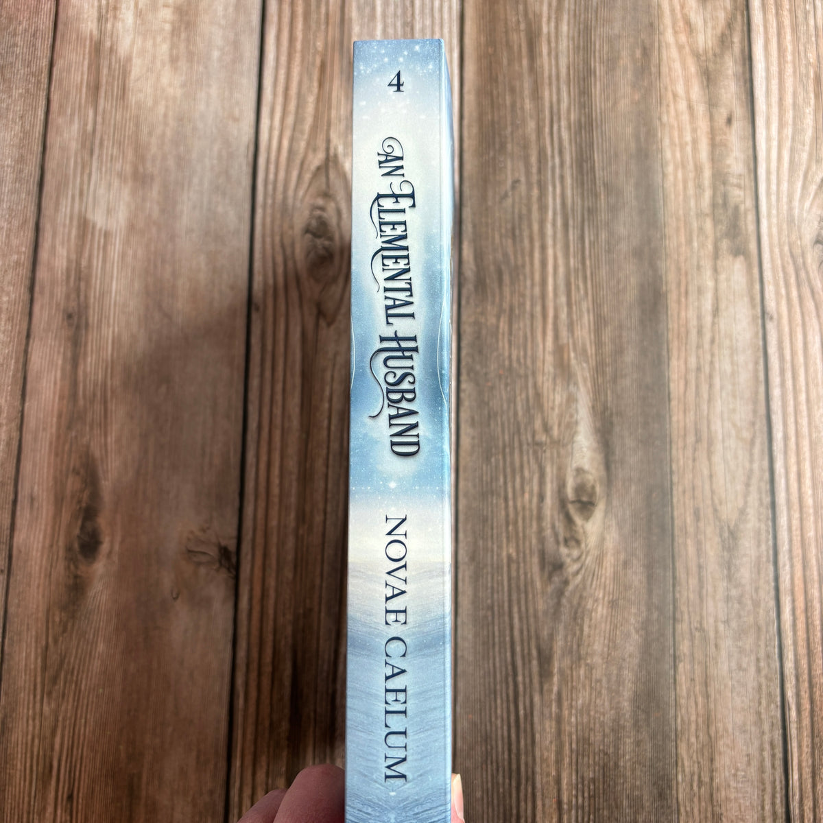 Spine of the paperback "An Elemental Husband: The King's Weaver Book 4" by Novae Caelum, held in hand against a wood background, showing the "4" volume number and icy blue cover design.