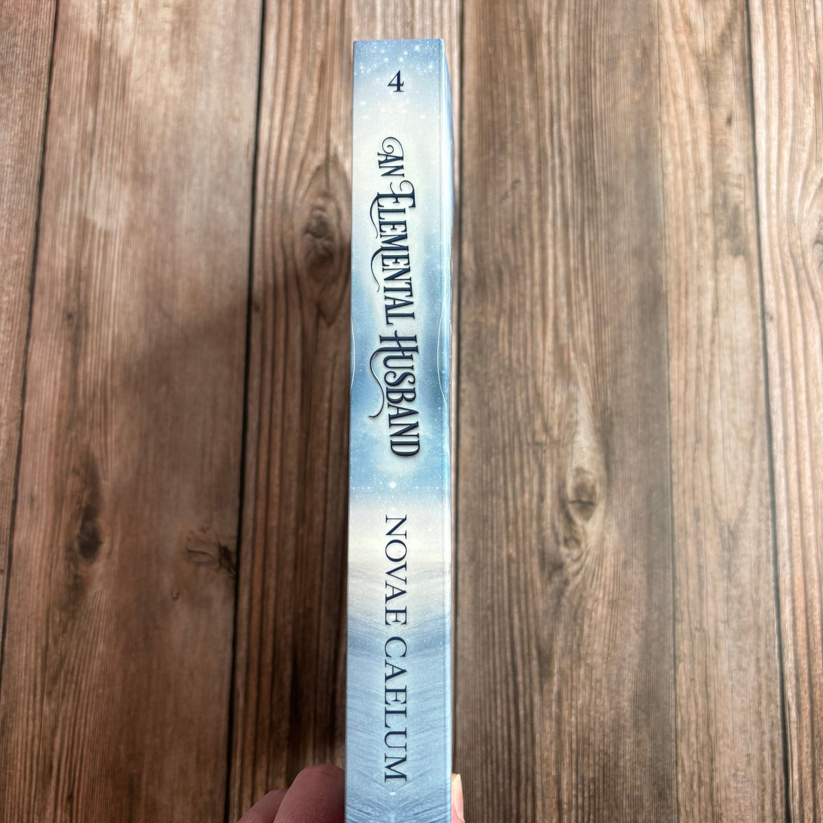 Spine of the unsigned paperback "An Elemental Husband: The King's Weaver Book 4" by Novae Caelum, held in hand against a wood background, showing the "4" volume number and icy blue cover design.
