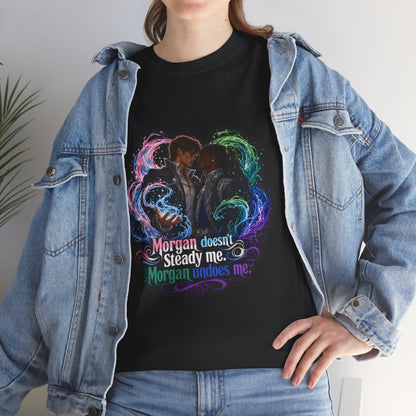 Black unisex t-shirt worn by a woman with long wavy brown hair, layered under an open denim jacket, printed with fantasy art of two men embracing in swirling magic and the quote "Morgan doesn't steady me, Morgan undoes me," close-up torso view.