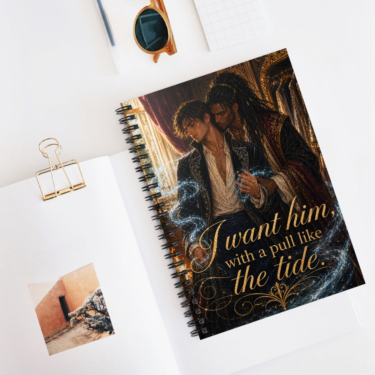 Spiral notebook open on a desk beside sunglasses, a pencil, and a small photo, showing the two men's embrace on the cover and the gold quote "I want him, with a pull like the tide."