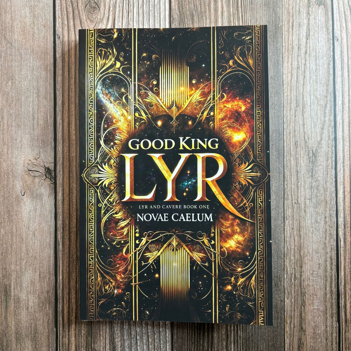 Photograph of Good King Lyr paperback by Novae Caelum standing on a wood-plank surface, its cover a gold-foil ornamental border framing a starfield and nebula.