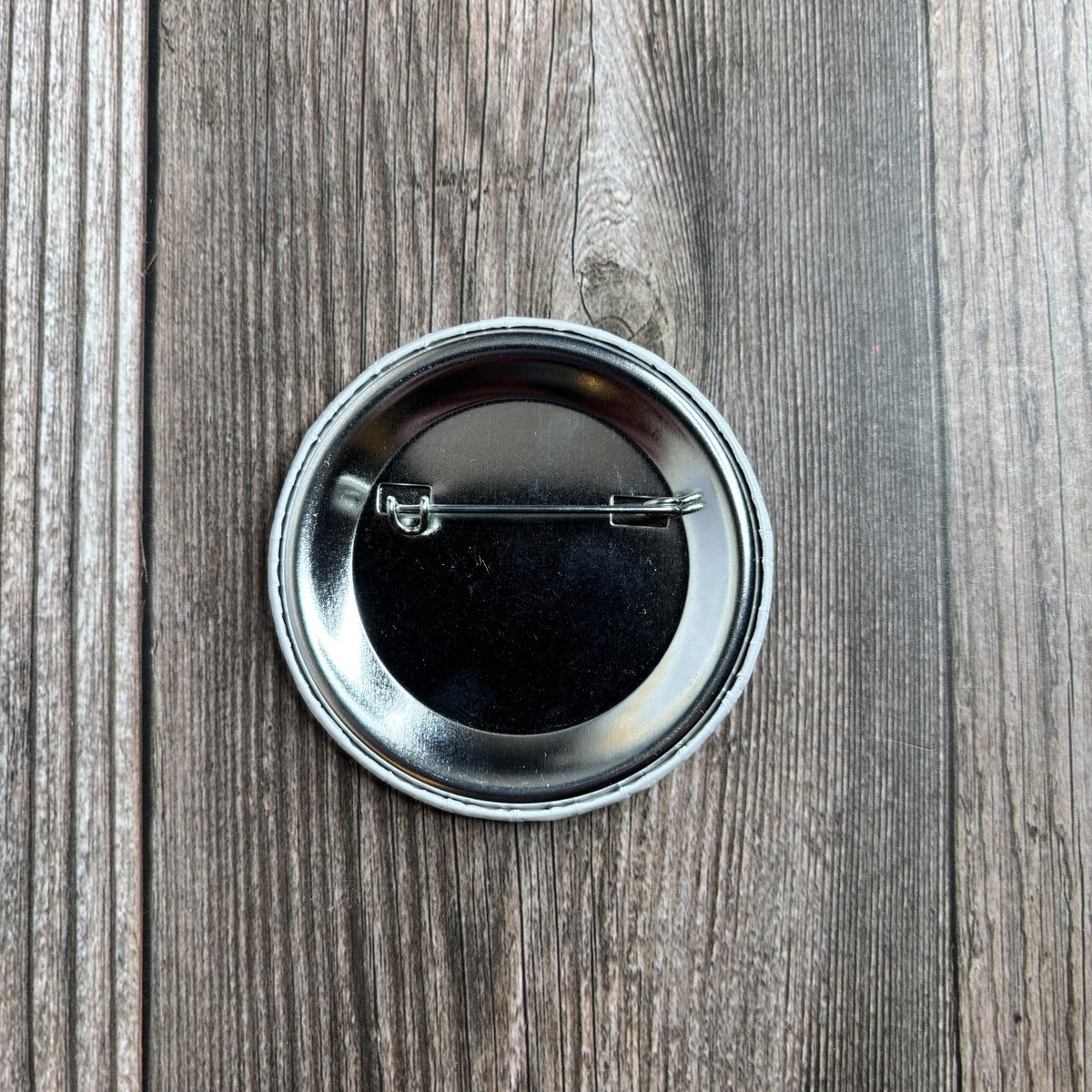 Back of the round button pin on a wood-grain surface, showing the metal pin-back fastener.