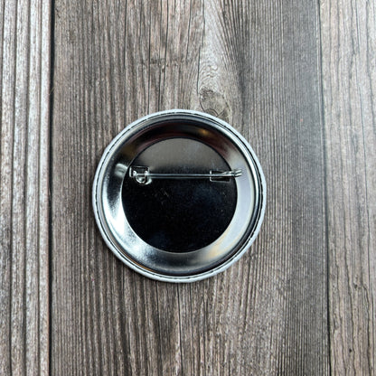 Back of the round button pin on a wood-grain surface, showing the metal pin-back fastener.