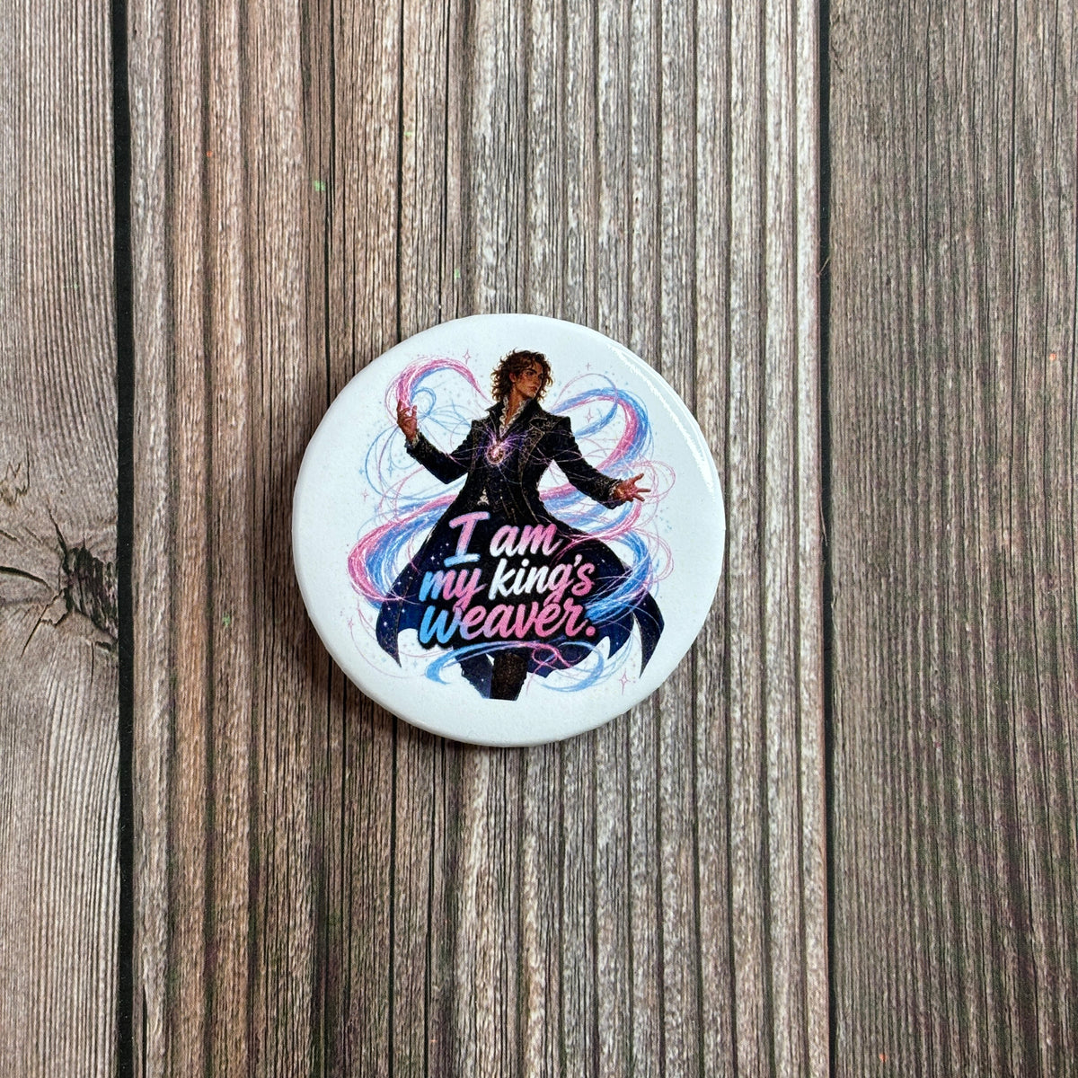 Round button pin lying on a wood-grain surface, illustration of a wavy-haired man in a black coat with swirling blue and pink magic, text "I am my king's weaver."