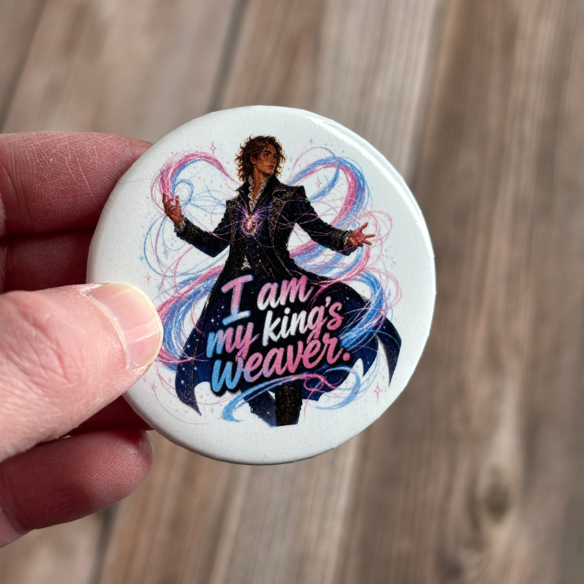 Round button pin held between fingers, illustration of a wavy-haired man in a black ornate coat with swirling blue and pink magic, text "I am my king's weaver."