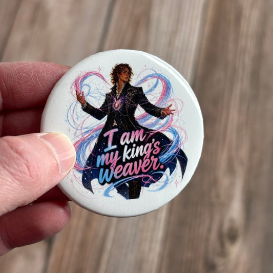 Round button pin held between fingers, illustration of a wavy-haired man in a black ornate coat with swirling blue and pink magic, text "I am my king's weaver."