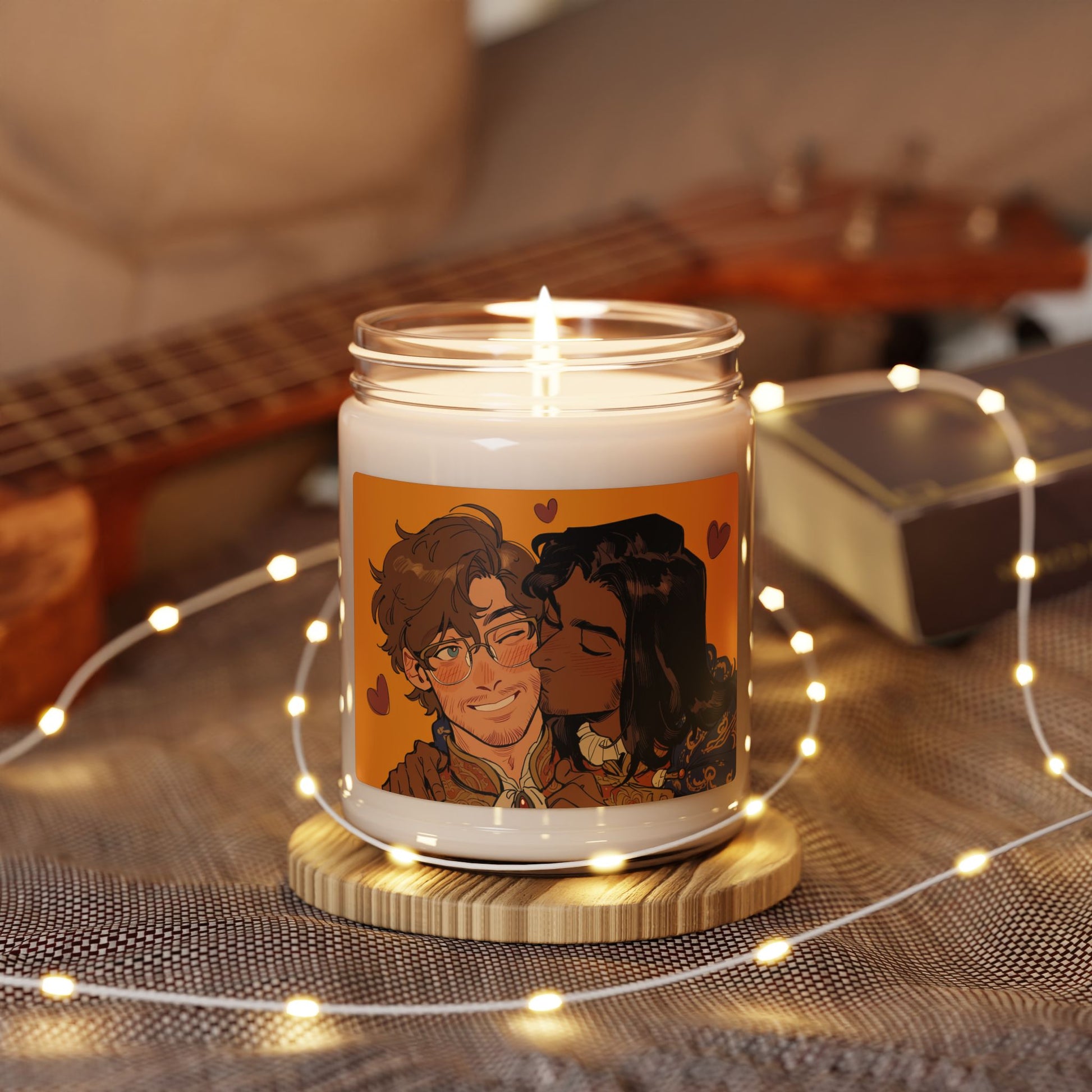 Lit 9oz soy candle on a wooden coaster surrounded by string lights, label art of a curly-haired man being kissed on the cheek by his dark-haired partner.