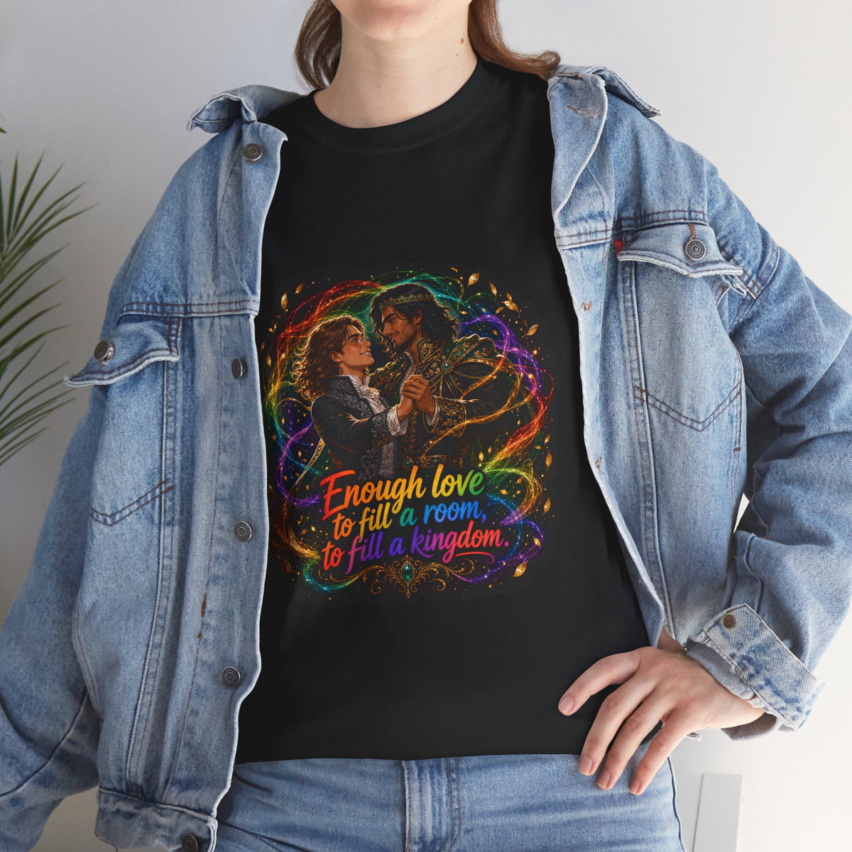 Black unisex t-shirt worn by a woman with long wavy brown hair, layered under an open denim jacket, printed with fantasy art of two men in formal attire dancing, wrapped in swirling rainbow magic and gold leaves, and the quote "Enough love to fill a room, to fill a kingdom."