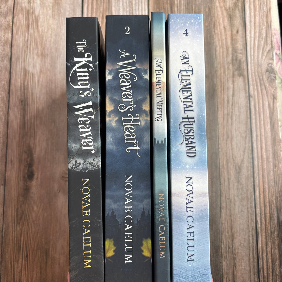 A hand holds four numbered hardcover spines from The King's Weaver series by Novae Caelum—The King's Weaver, A Weaver's Heart, An Elemental Meeting, and An Elemental Husband—against a wood background.