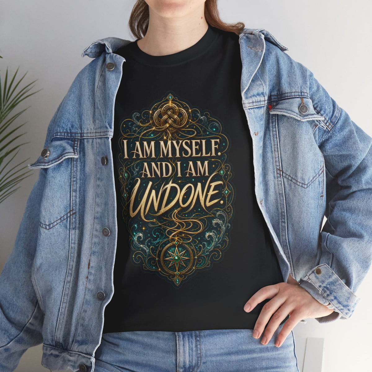 Black unisex t-shirt worn by a woman with long brown hair, layered under an open denim jacket, printed with a gold Celtic knot design and the quote "I am myself. And I am undone."