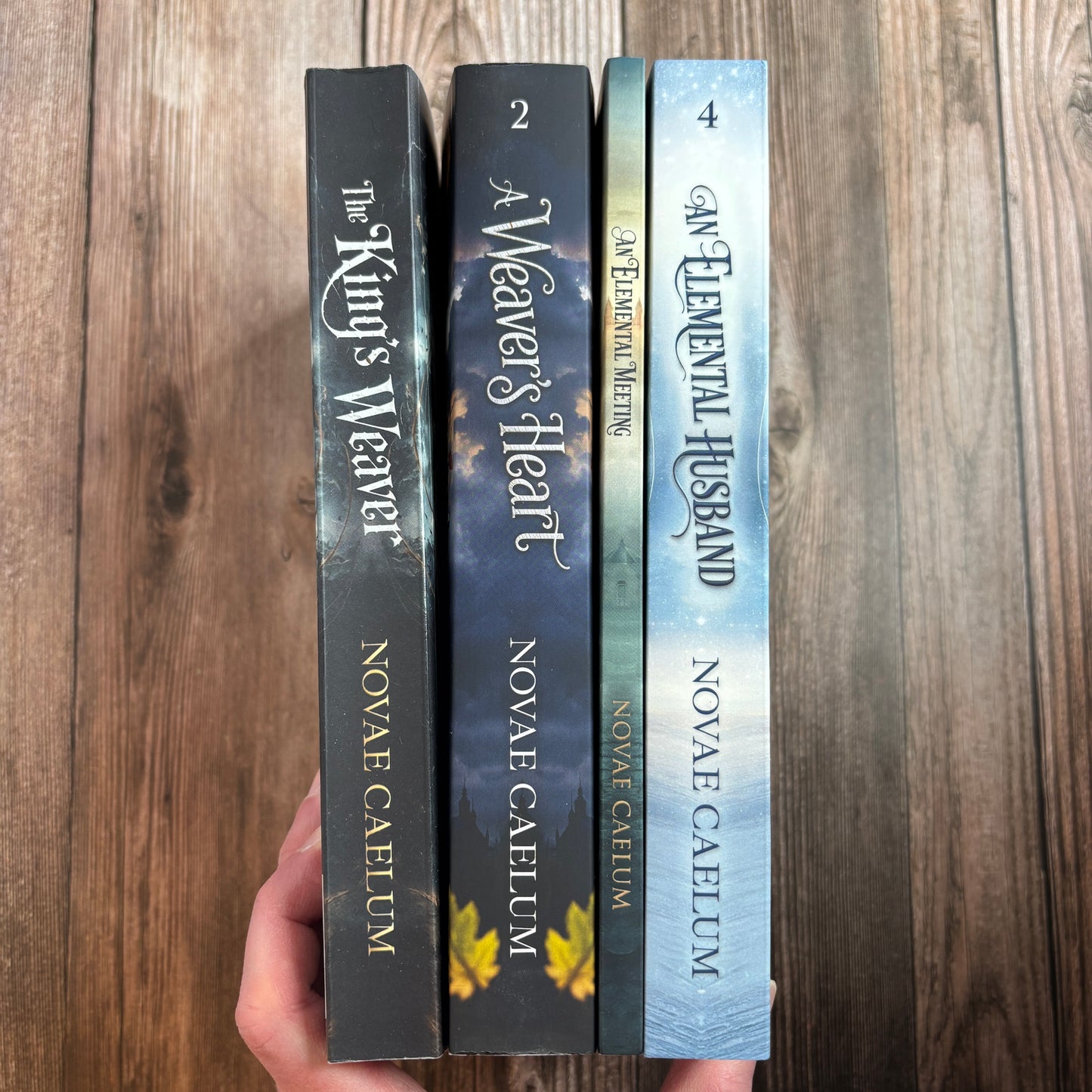 A hand holds four numbered paperback spines from The King's Weaver series by Novae Caelum—The King's Weaver, A Weaver's Heart, An Elemental Meeting, and An Elemental Husband—against a wood background.