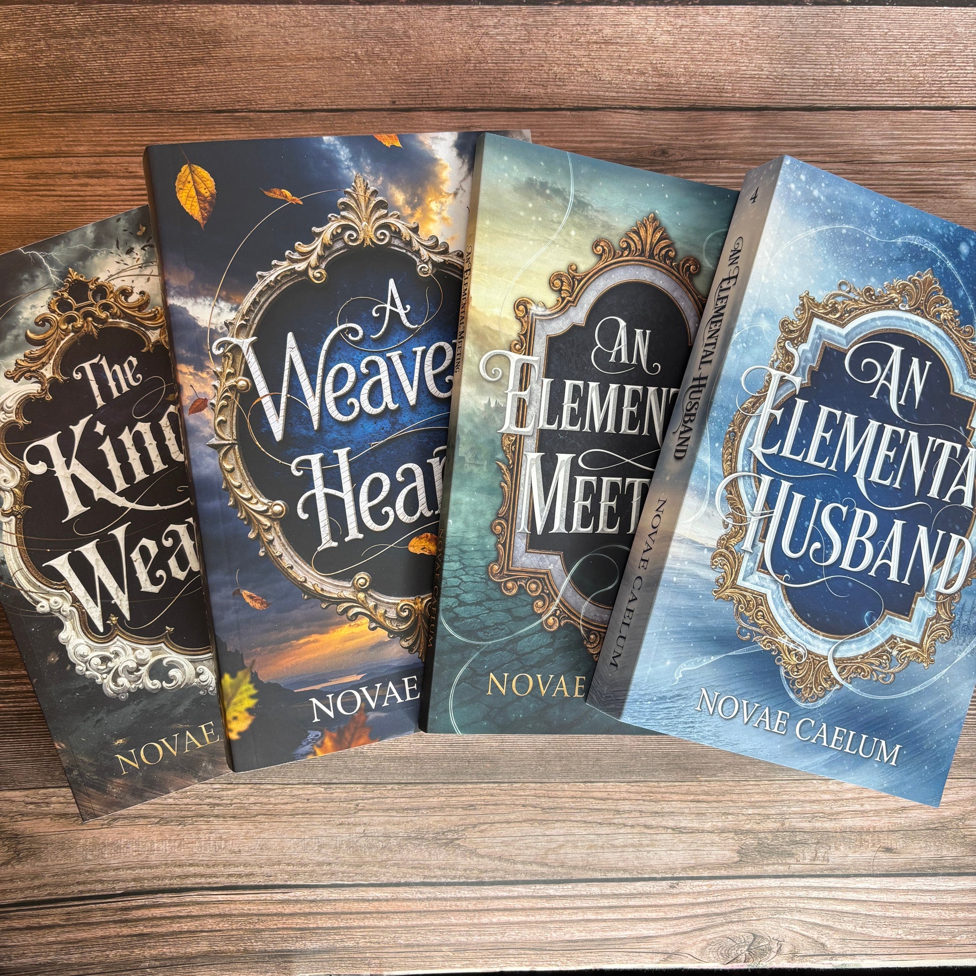Four fanned paperbacks of The King's Weaver series by Novae Caelum on a wood-plank surface - The King's Weaver, A Weaver's Heart, An Elemental Meeting, and An Elemental Husband - each with an ornate gold-framed cover.