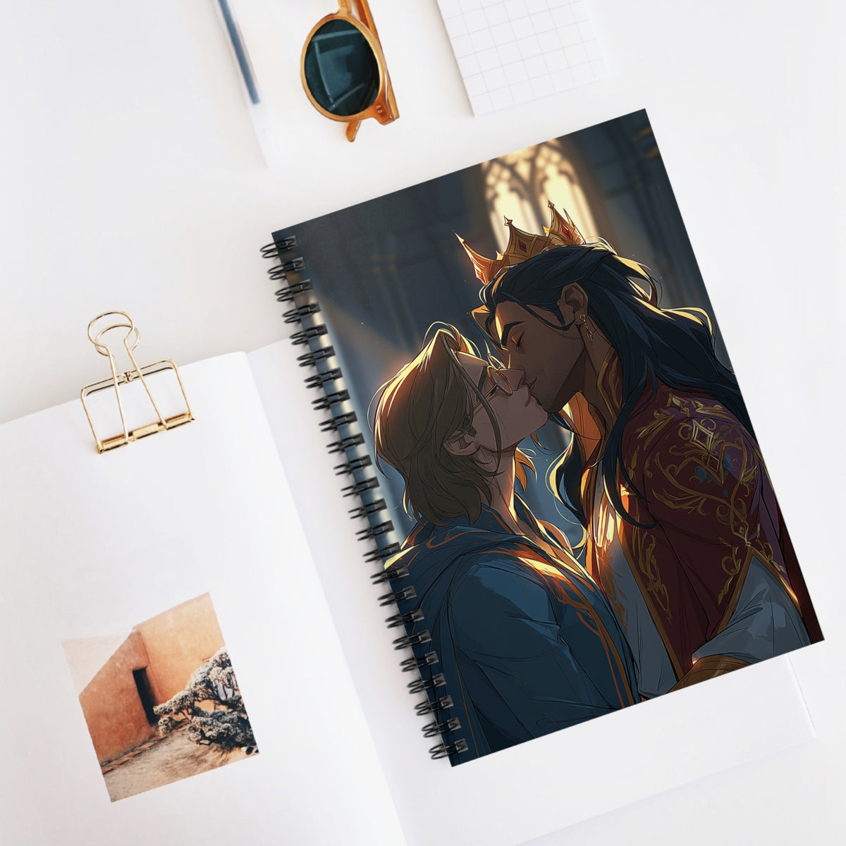 Spiral notebook open on a desk beside sunglasses, a pencil, and a small photo, showing the crowned couple kissing on the cover in golden cathedral light.