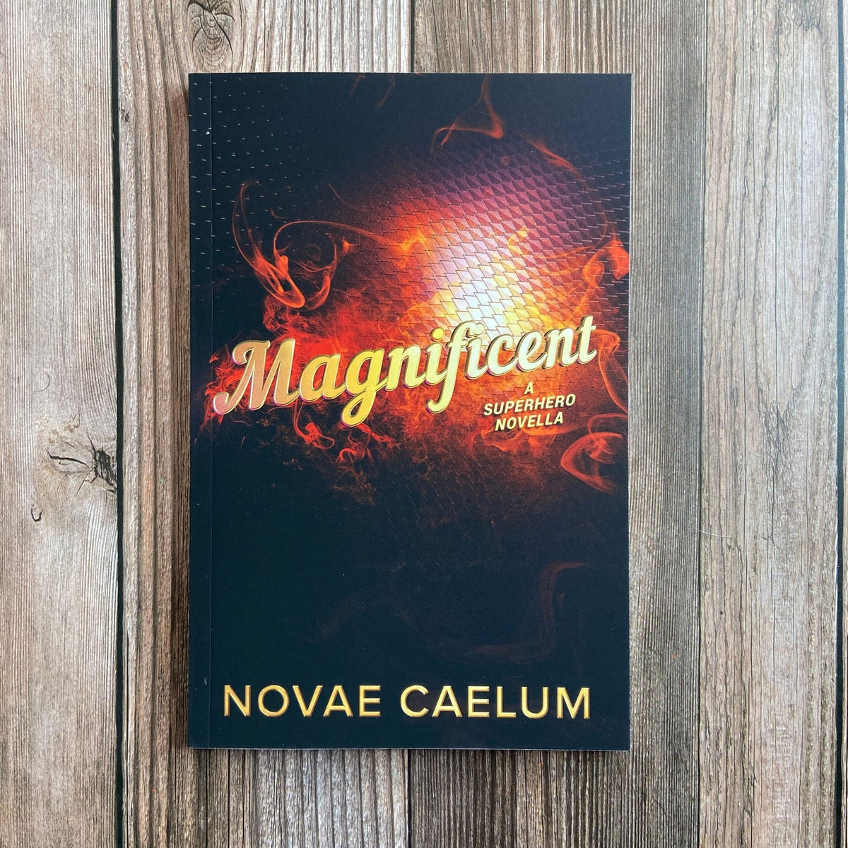 Photograph of Magnificent: A Superhero Novella paperback by Novae Caelum standing on a wood-plank surface, its cover a fiery orange-and-black swirl behind gold script title text.