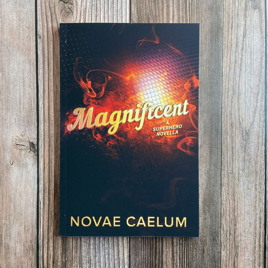 Photograph of Magnificent: A Superhero Novella paperback by Novae Caelum standing on a wood-plank surface, its cover a fiery orange-and-black swirl behind gold script title text.