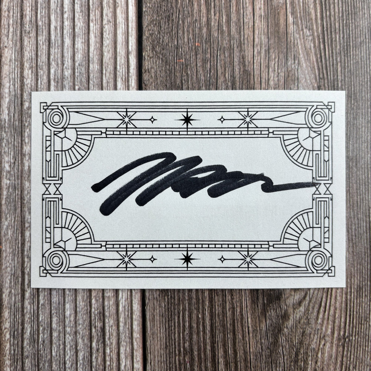 Rectangular signed bookplate with an ornate art-deco star-and-scroll border and a handwritten black-ink signature across the center, resting on a wood-grain surface.