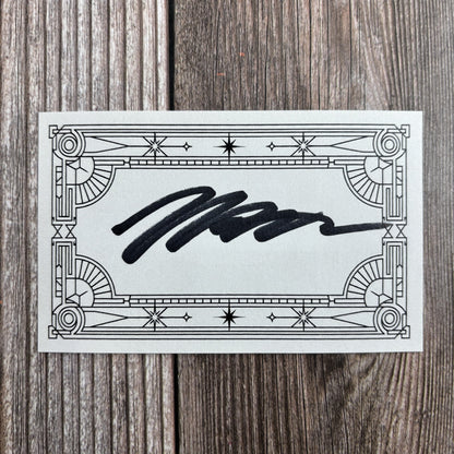 Rectangular signed bookplate with an ornate art-deco star-and-scroll border and a handwritten black-ink signature across the center, resting on a wood-grain surface.