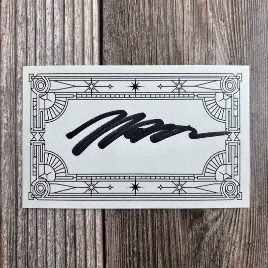 Rectangular signed bookplate with an ornate art-deco star-and-scroll border and a handwritten black-ink signature across the center, resting on a wood-grain surface.