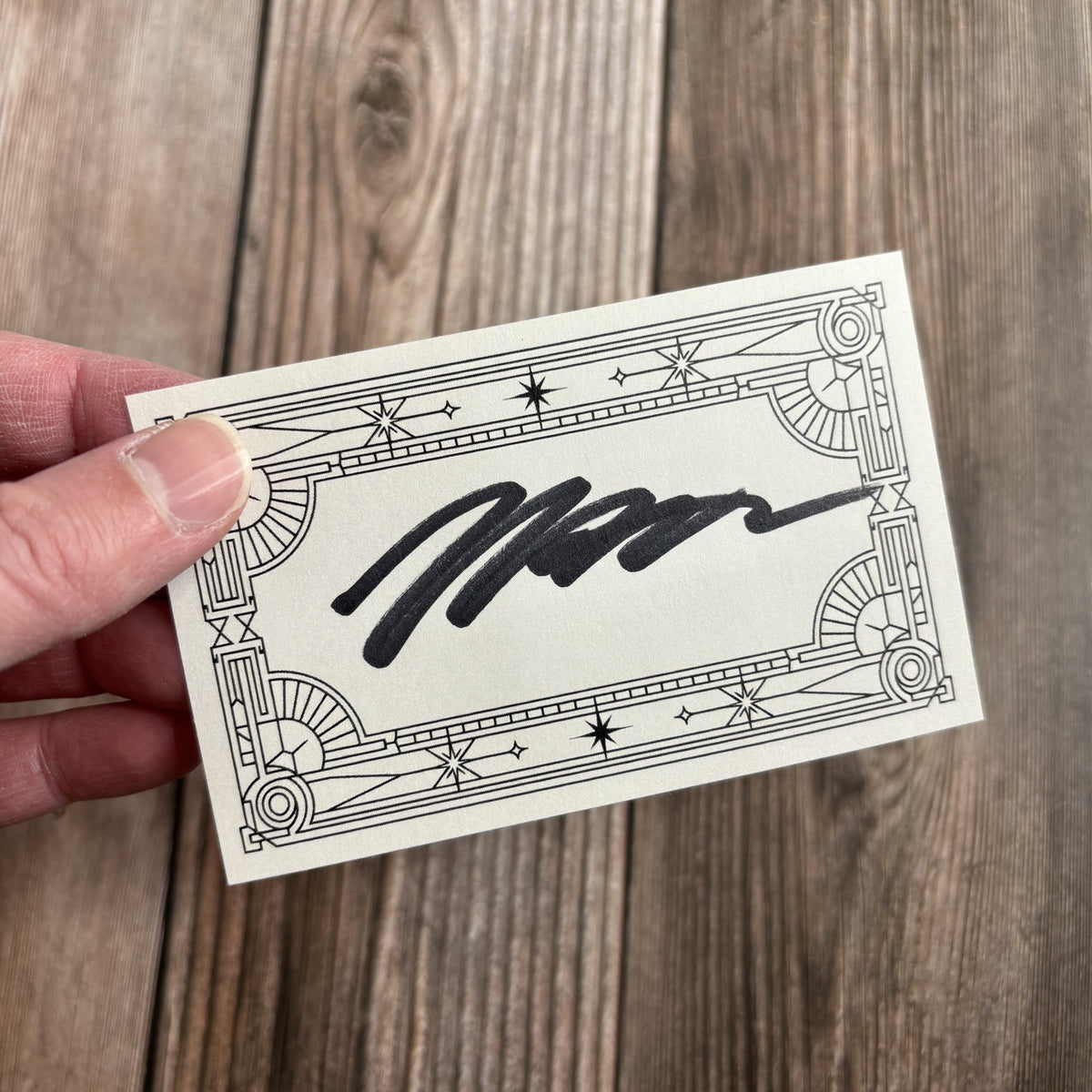 A hand holding the signed bookplate at an angle, showing its art-deco border and handwritten black-ink signature, against a wood-grain background.