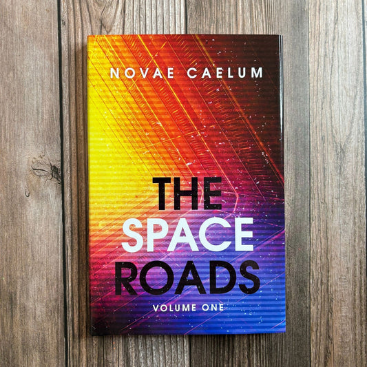 Photograph of The Space Roads: Volume One paperback by Novae Caelum standing on a wood-plank surface, its cover a diagonal orange-to-blue gradient with bold title text.