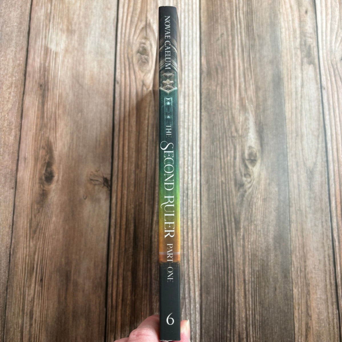 Spine of "The Second Ruler Part One: The Stars and Green Magics Book 6" paperback by Novae Caelum, held in hand against a wood-plank background, showing the title, series name, and book number 6 (Spine Cover Paperback).