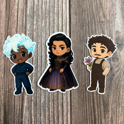 Three chibi character stickers from "The Second Ruler Part Two: The Stars and Green Magics Book 7" on wood: a blue-haired man in blue, a dark-haired woman in a purple gown, and a curly-haired man holding a purple flower.