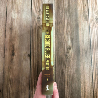 Spine of "The Second Ruler Part Two: The Stars and Green Magics Book 7" paperback by Novae Caelum, held in hand against a wood-plank background, showing the title, series name, and book number 7 (Spine Cover Paperback).