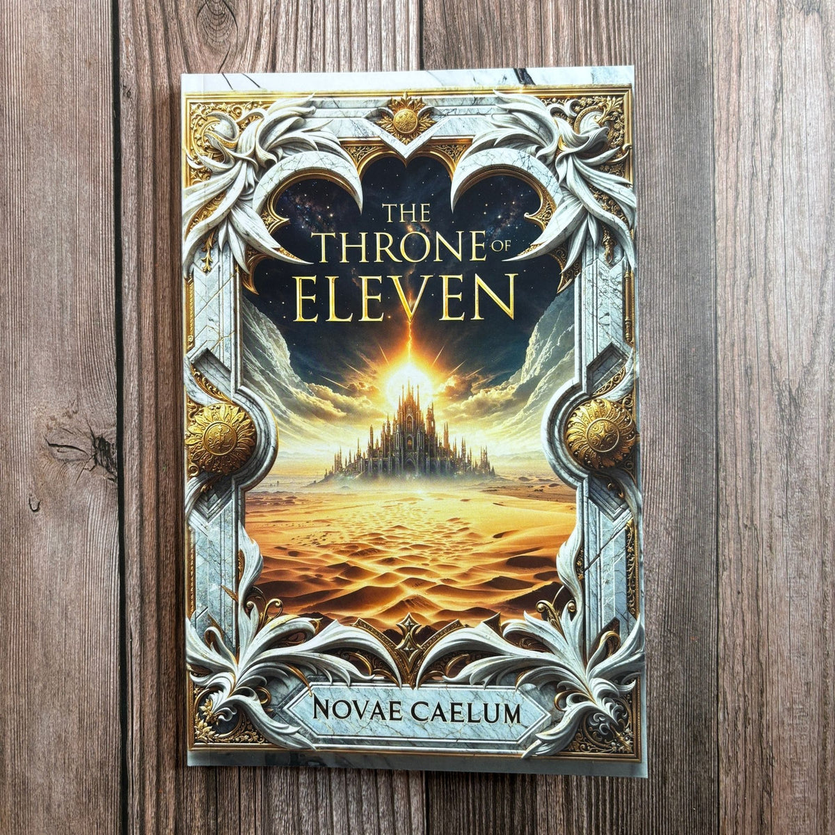 Photograph of The Throne of Eleven paperback by Novae Caelum standing on a wood-plank surface, its cover showing an ornate marble-and-gold frame around a glowing desert palace.