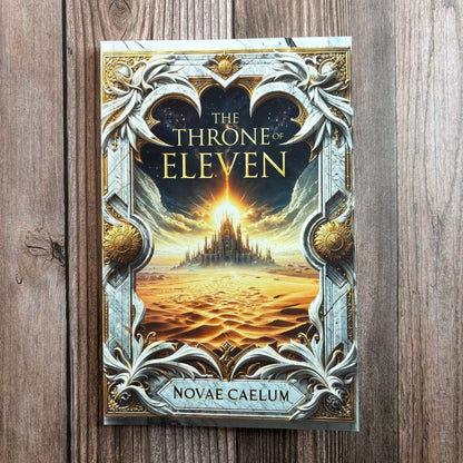 Photograph of The Throne of Eleven paperback by Novae Caelum standing on a wood-plank surface, its cover showing an ornate marble-and-gold frame around a glowing desert palace.