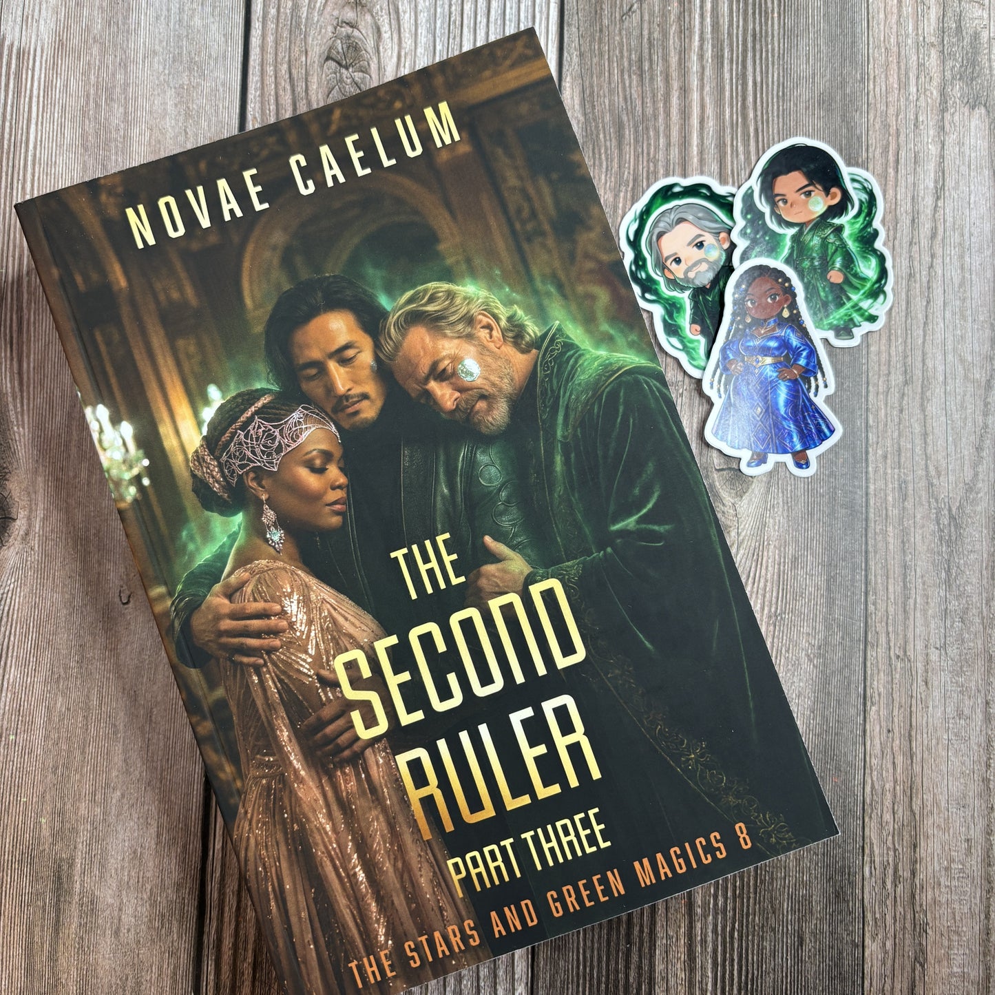 Character-art edition paperback of "The Second Ruler: Part Three" by Novae Caelum lying at an angle on weathered wood with three chibi character stickers beside it.