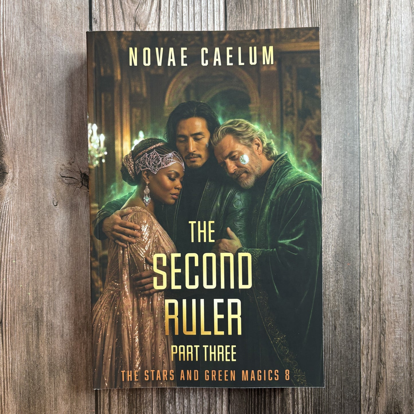 Front cover of the character-art edition of "The Second Ruler: Part Three — The Stars and Green Magics Book 8" by Novae Caelum, paperback on weathered wood showing three people embracing in a gilded, green-lit hall: a Black woman in a beaded headpiece and rose-gold gown, a dark-haired man of East Asian descent, and a white-bearded man in green velvet robes.