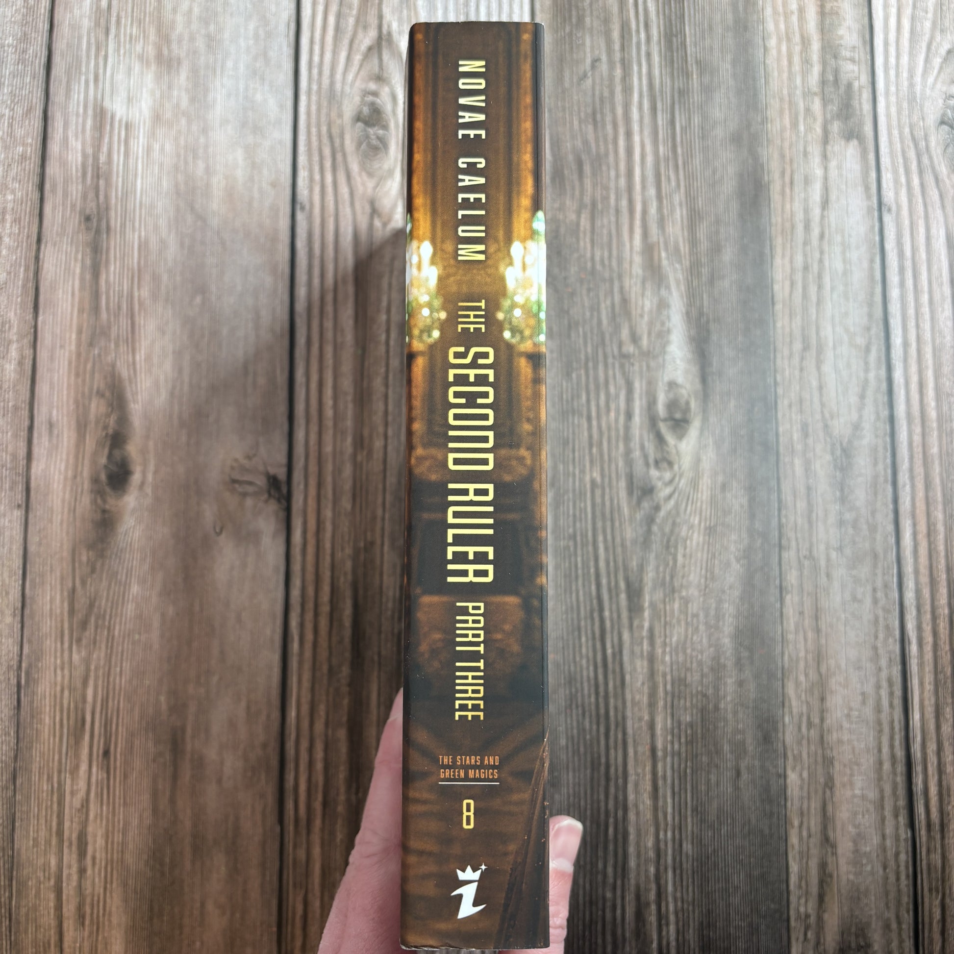 Spine of the character-art edition of "The Second Ruler: Part Three" paperback by Novae Caelum held in hand, gold lettering over dark gilded hall art, with the series name, book number 8, and the Ialorius Press logo at the base.