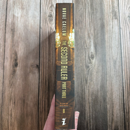 Spine of the character-art edition of "The Second Ruler: Part Three" paperback by Novae Caelum held in hand, gold lettering over dark gilded hall art, with the series name, book number 8, and the Ialorius Press logo at the base.