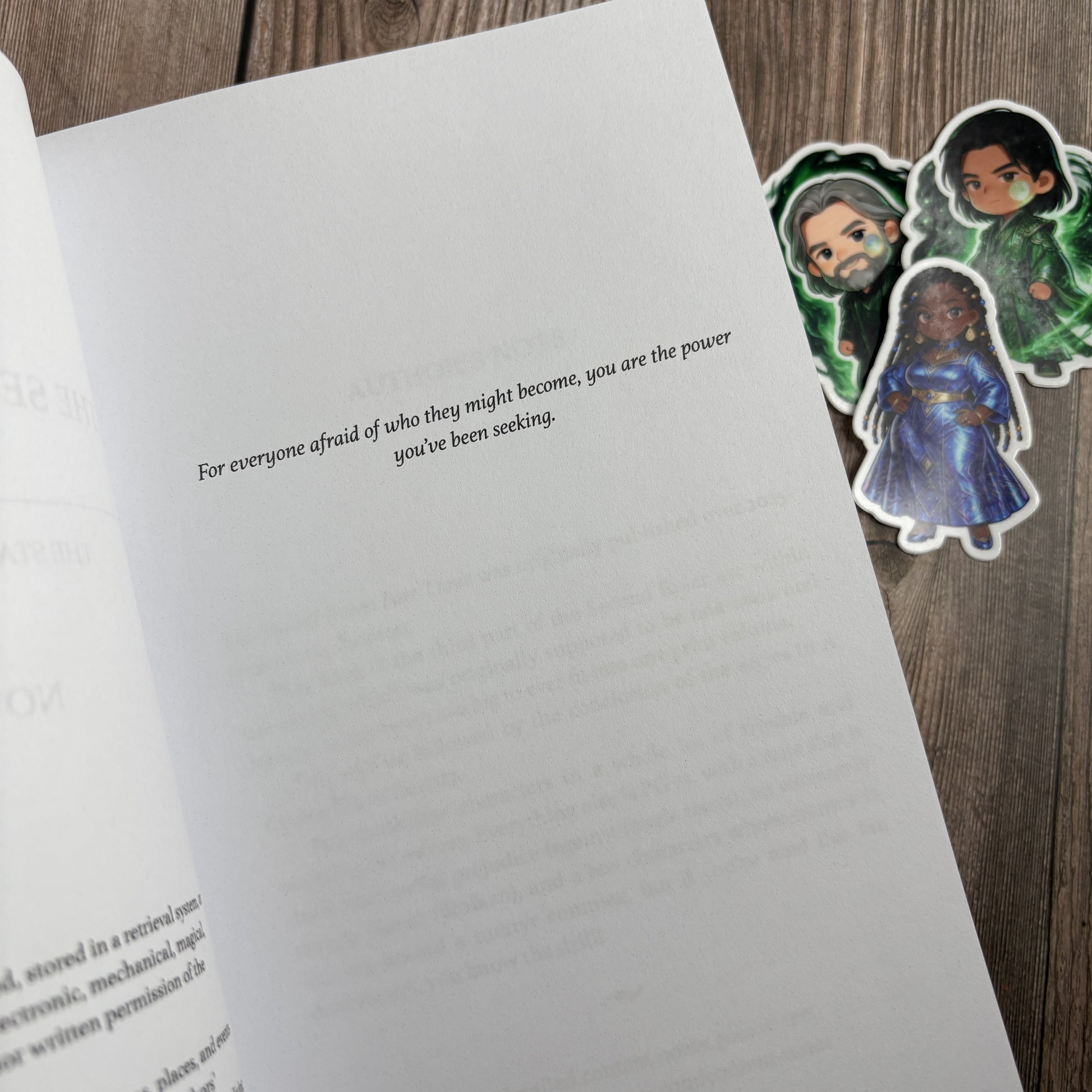 Open dedication page of "The Second Ruler: Part Three" by Novae Caelum reading "For everyone afraid of who they might become, you are the power you've been seeking," with three chibi character stickers on the wood beside the book.