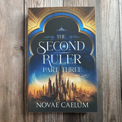 Front cover of "The Second Ruler: Part Three — The Stars and Green Magics Book 8" by Novae Caelum, paperback lying on weathered grey wood, showing an ornate blue-and-gold archway framing a golden spired city beneath a starry sky.