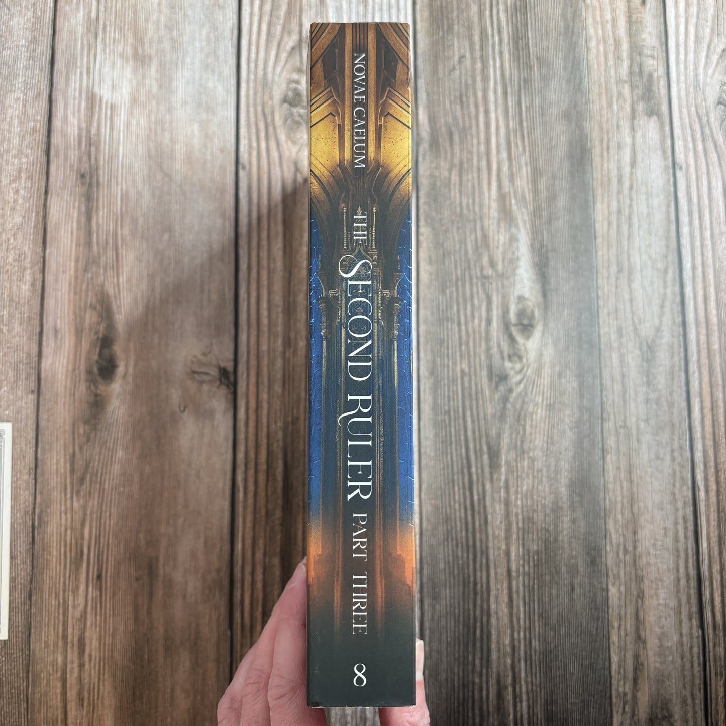 Spine of "The Second Ruler: Part Three" paperback by Novae Caelum held in hand against weathered wood, reading "Novae Caelum — The Second Ruler, Part Three" with the series number 8 at the base.