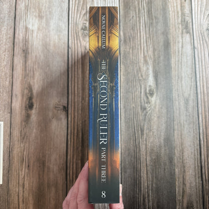Spine of "The Second Ruler: Part Three" paperback by Novae Caelum held in hand against weathered wood, reading "Novae Caelum — The Second Ruler, Part Three" with the series number 8 at the base.