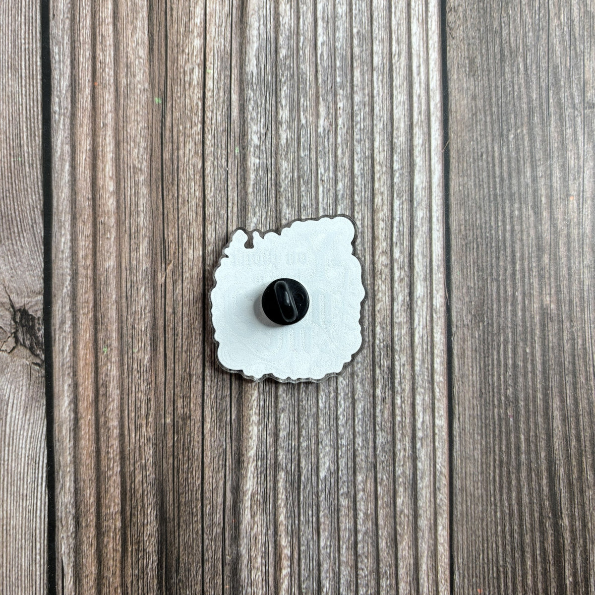 Back of the die-cut acrylic pin on a wood-grain surface, showing its black rubber pin backing.