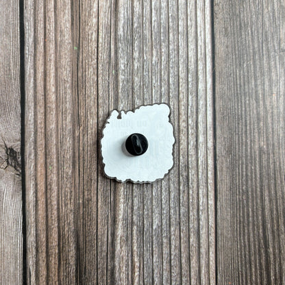 Back of the die-cut acrylic pin on a wood-grain surface, showing its black rubber pin backing.