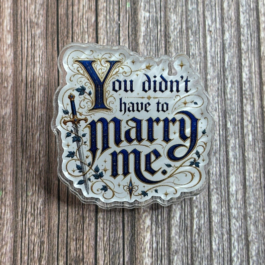 Die-cut holographic acrylic pin on a wood-grain surface, illuminated-manuscript-style navy and gold text "You didn't have to marry me." beside an ivy-wrapped sword.