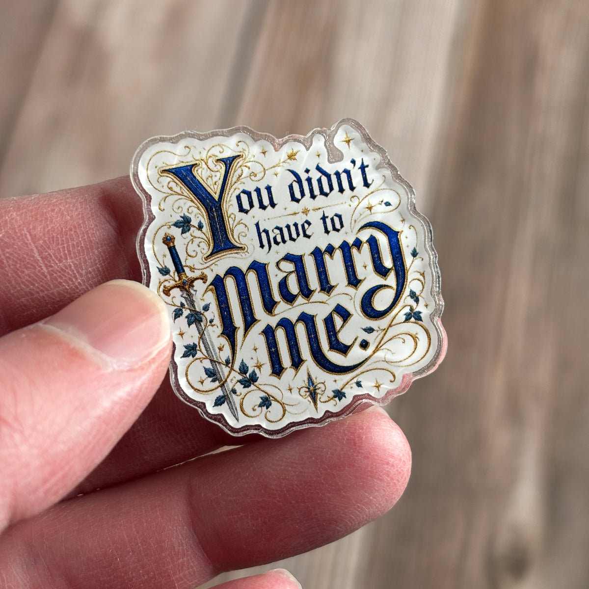Die-cut holographic acrylic pin held between fingers, illuminated-manuscript-style navy and gold text "You didn't have to marry me." beside an ivy-wrapped sword.